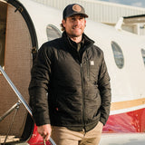 A man wearing the Flight Outfitters Airfoil Jacket, standing confidently in front of an aircraft.