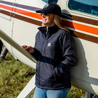 Woman wearing Flight Outfitters Women's Airfoil Insulated Jacket with hat, hands in pockets, in front of Cessna high-wing aircraft