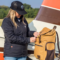 Woman wearing Flight Outfitters Women's Airfoil Insulated Jacket with hat, opening backpack in front of Cessna high-wing aircraft